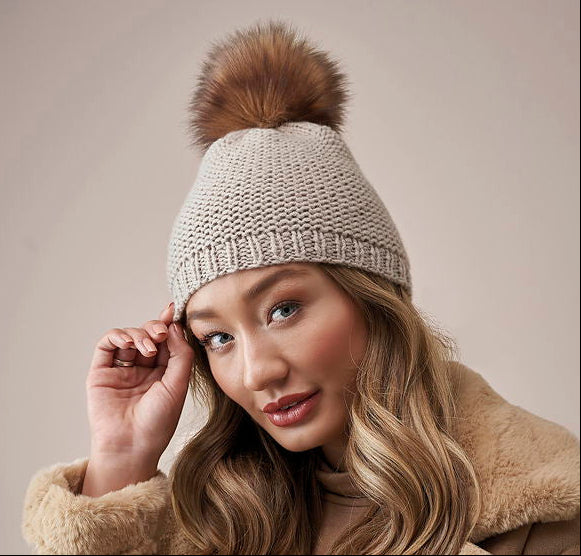 Woman wearing a beige knitted beanie with a brown fur pom-pom against a neutral background