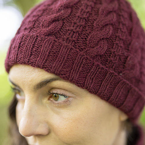 A close-up of a woman wearing a burgundy Darvel cable-knit beanie hat, with a soft green bokeh background.