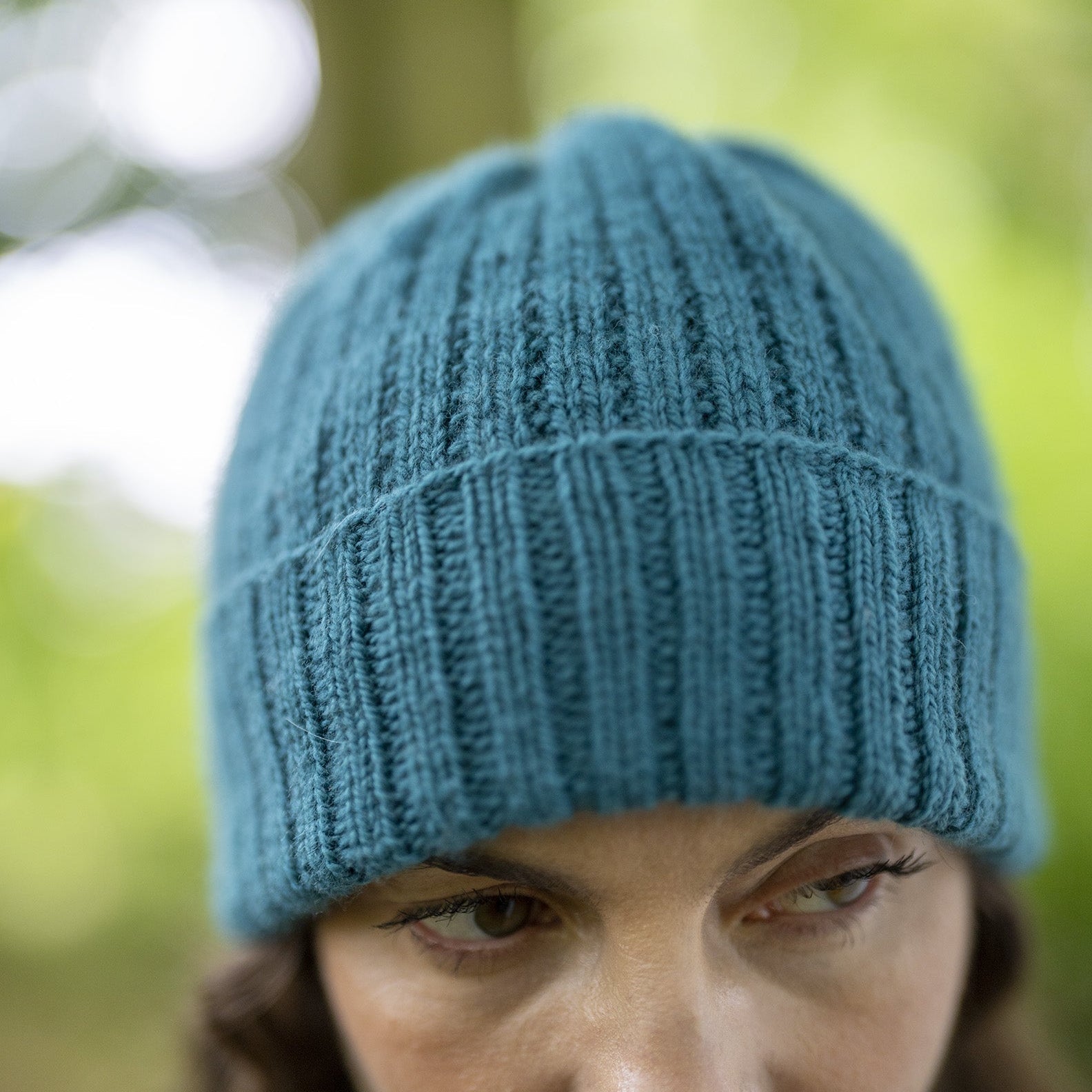 A close-up shot of a woman wearing a teal blue ribbed knit beanie hat, with a soft-focus outdoor background of green foliage.
