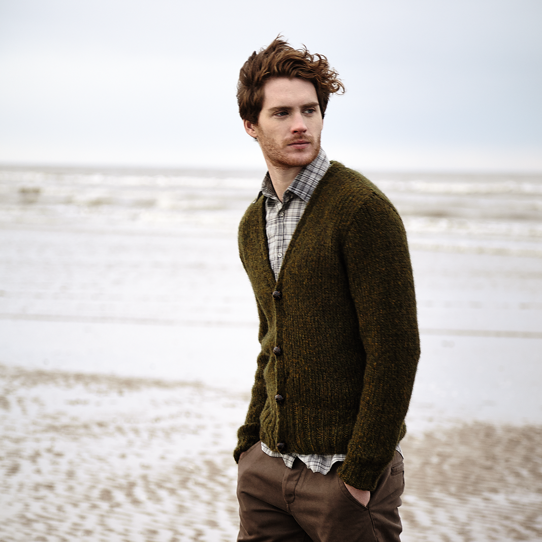 Man wearing a green Stour cardigan and brown pants standing on a beach.
