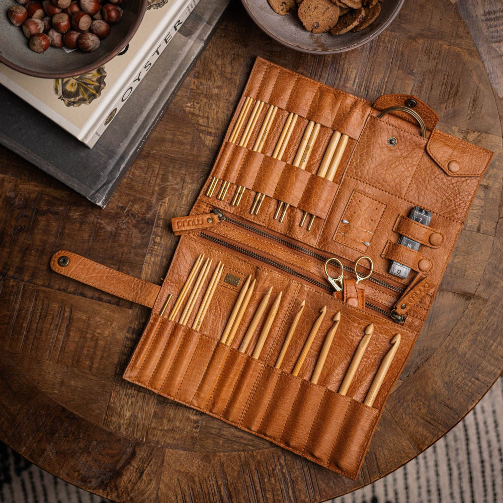 muud Billlie whisky coloured leather case for crochet hooks and knitting needles is displayed open on a table, revealing multiple compartments and pockets with various tools organized inside.
