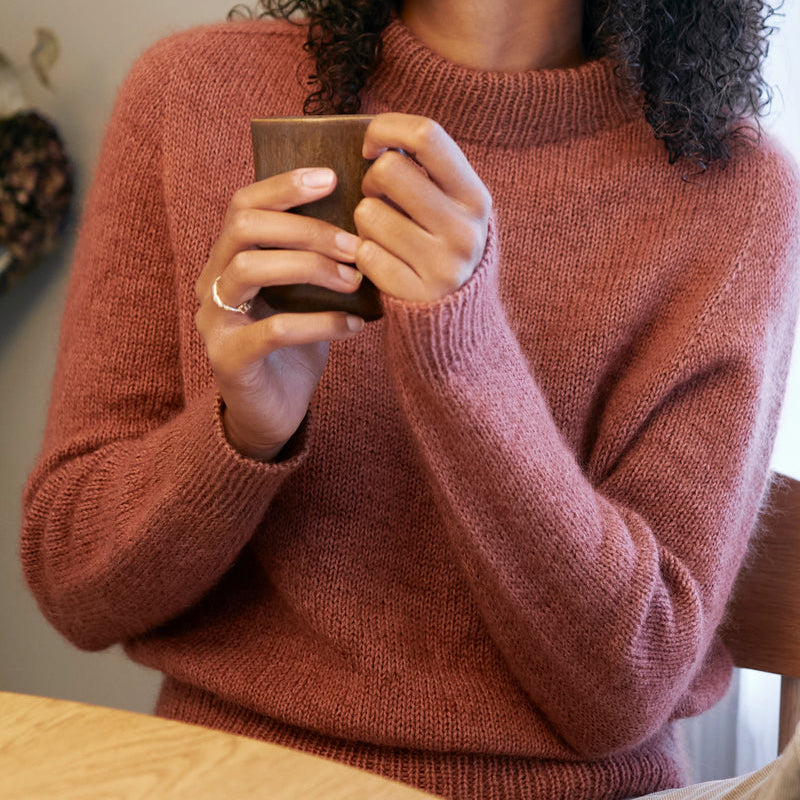 Person wearing a pink Knitting for Olive Carlie sweater holding a brown mug indoors.