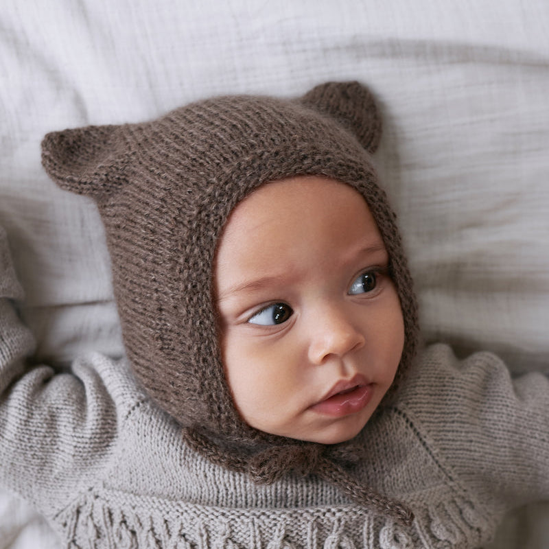 A baby wearing the Knitting for Olive Baby Bear Bonnet, showing the rounded ears and I-cord ties