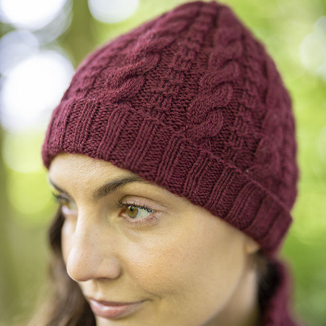 A close-up of a woman wearing a burgundy Darvel cable-knit beanie hat, with a soft green bokeh background.