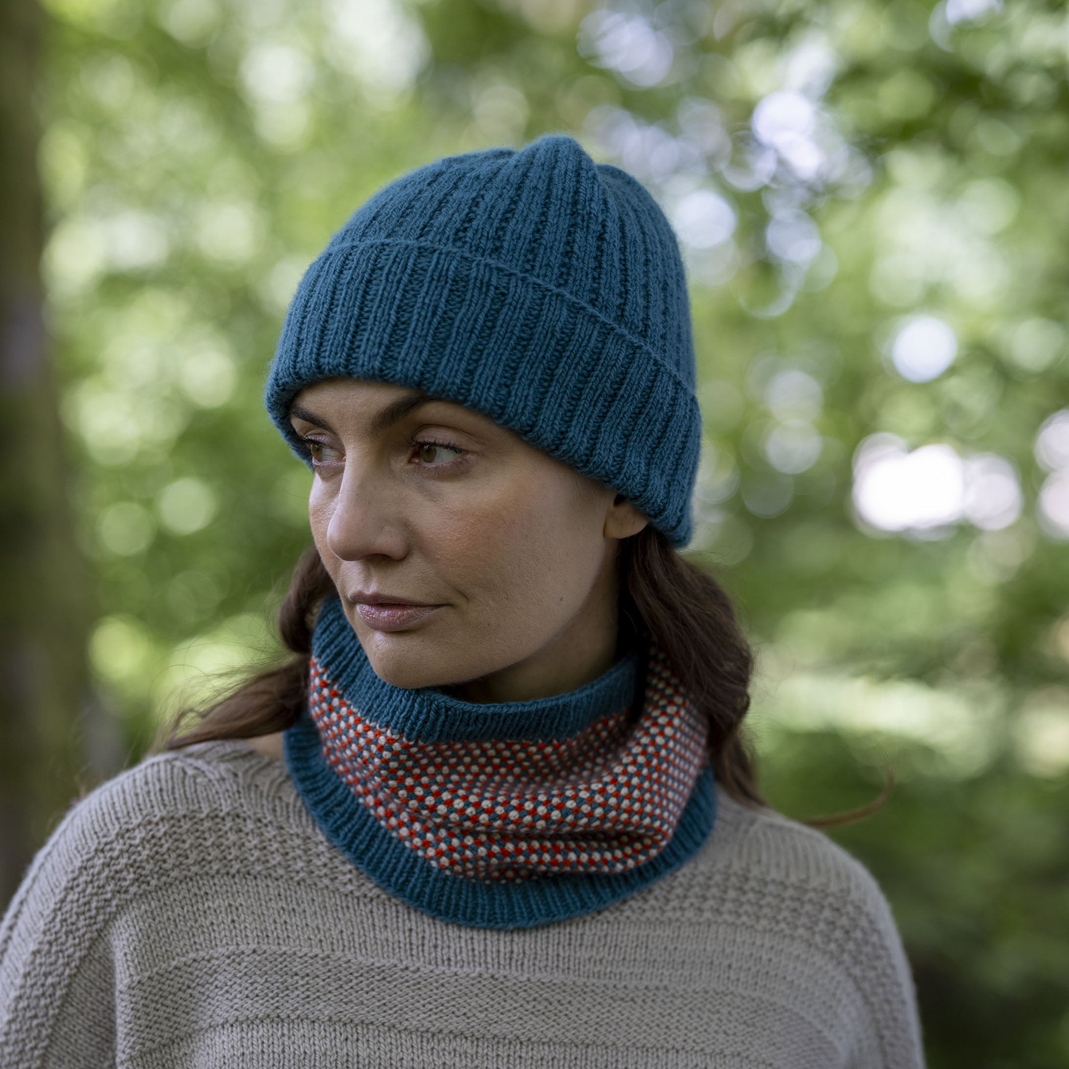 A woman wearing a teal blue ribbed knit beanie and a matching teal cowl neck warmer with a red and white patterned inner layer, photographed outdoors in a blurred green forest setting.